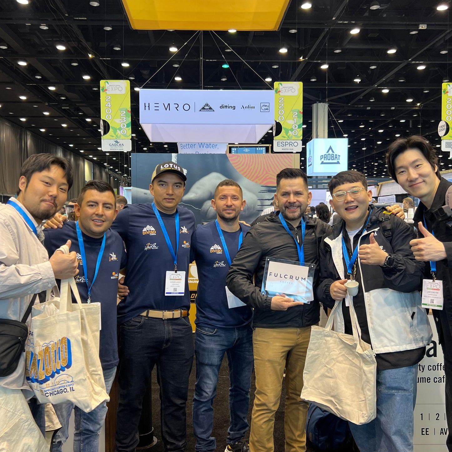 A group of seven men posing together at a convention or trade show booth, holding tote bags and standing in front of a display screen with the brand name HEMRO visible.