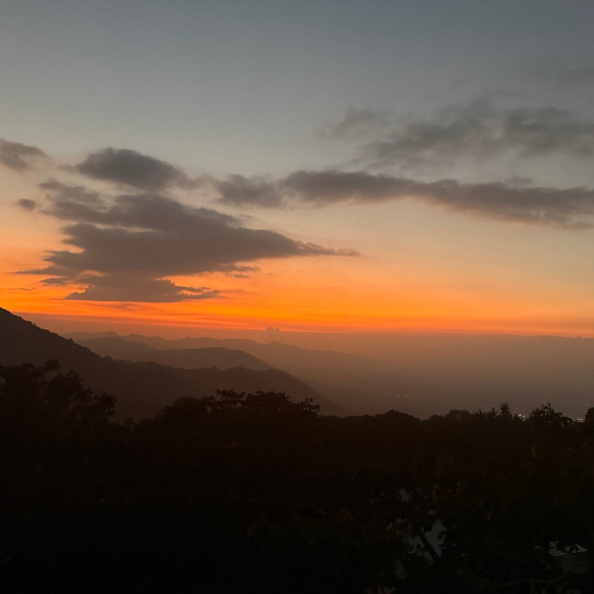 A scenic sunset landscape with layered mountain silhouettes in the foreground and a glowing orange horizon under a pale blue sky with scattered clouds.
