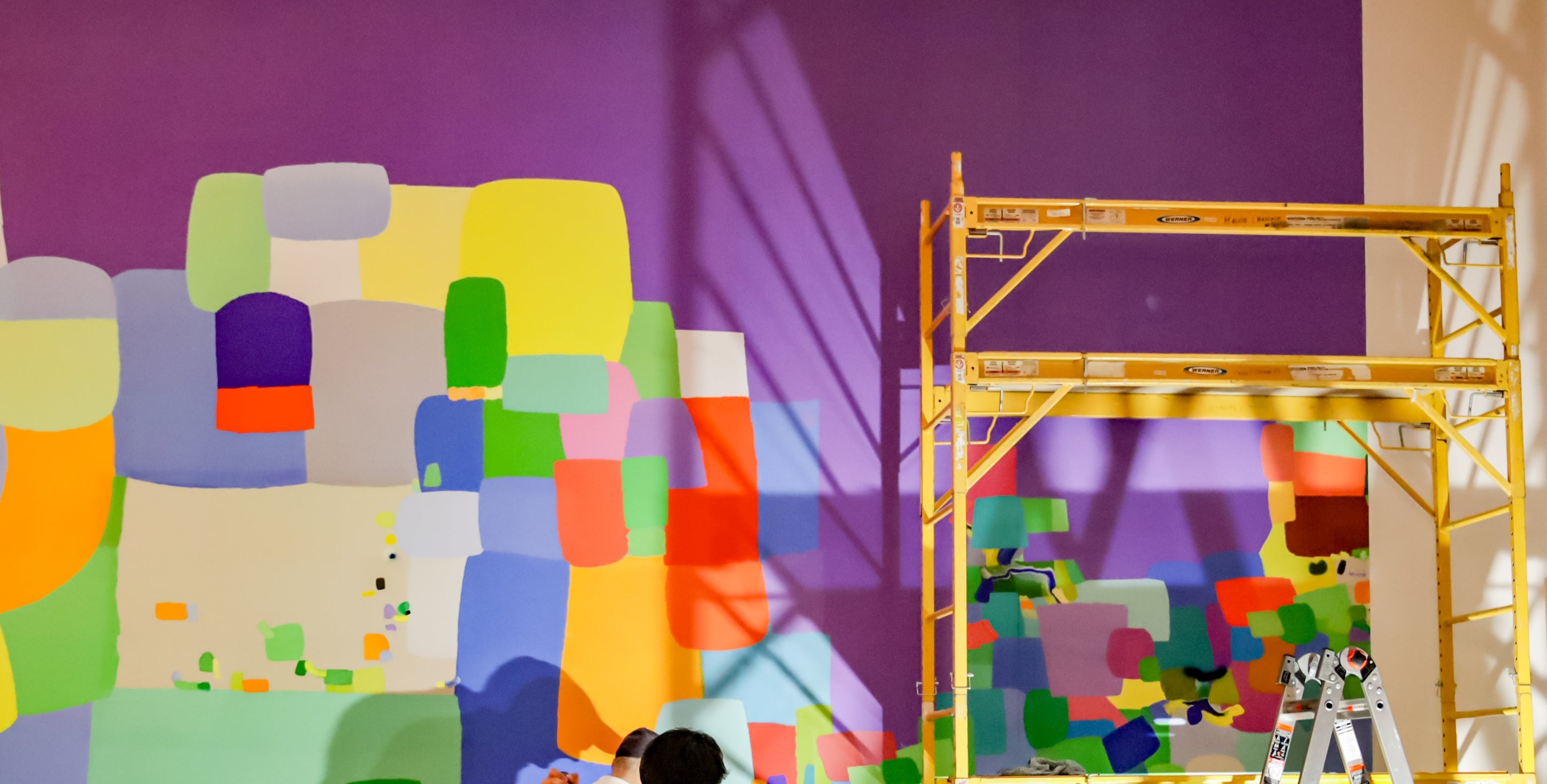 A group of painters working on a large colorful abstract wall mural with geometric blocks in yellow, green, blue, purple, orange, and red. A yellow scaffolding structure stands to the right against a purple wall.