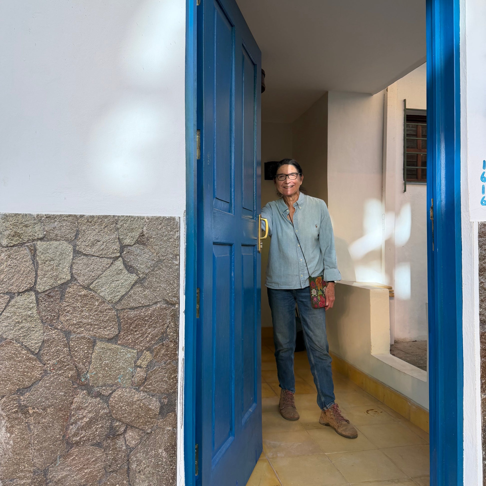 A woman wearing a light blue button-up shirt, blue jeans, and brown boots stands in a doorway with a bright blue frame, looking out onto a sunlit tiled patio. The wall on the left features a textured stone pattern.